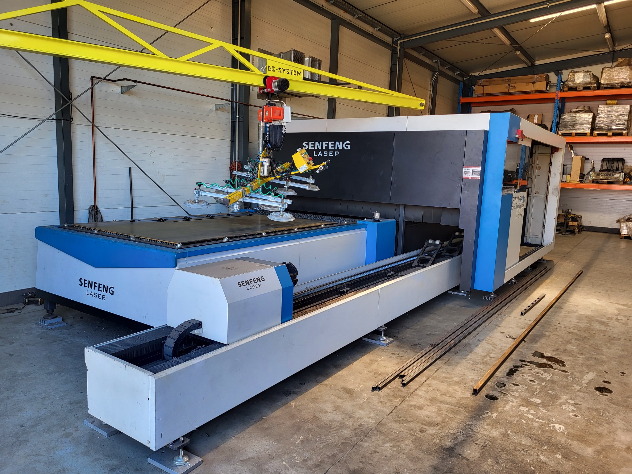 Laserowa maszyna CNC Senfeng z żółtym suwnicą i przyssawkami, gotowa do obróbki metalu w hali produkcyjnej. Widoczne profile metalowe na podłodze.