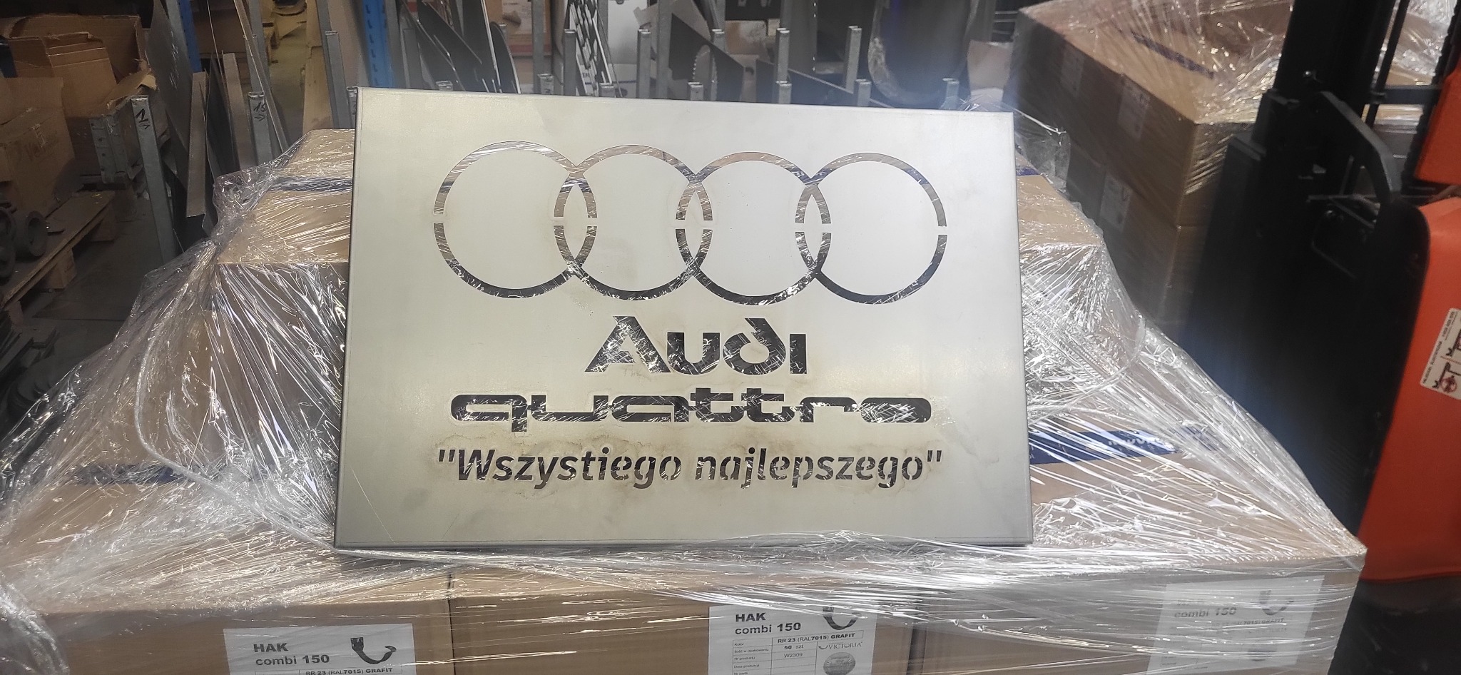 Metalowa tablica z wyciętym laserowo logo Audi Quattro i napisem 'Wszystkiego najlepszego', leżąca na palecie zafoliowanych kartonów w magazynie.