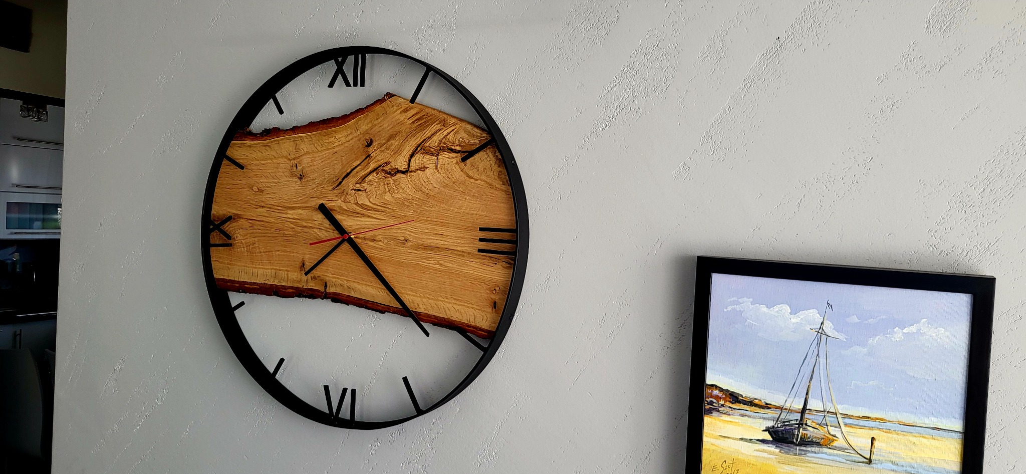 Oryginalny zegar ścienny z plastra drewna w czarnej, metalowej ramie, z czerwonymi wskazówkami, wiszący na teksturowanej, jasnej ścianie obok obrazu z motywem morskim.