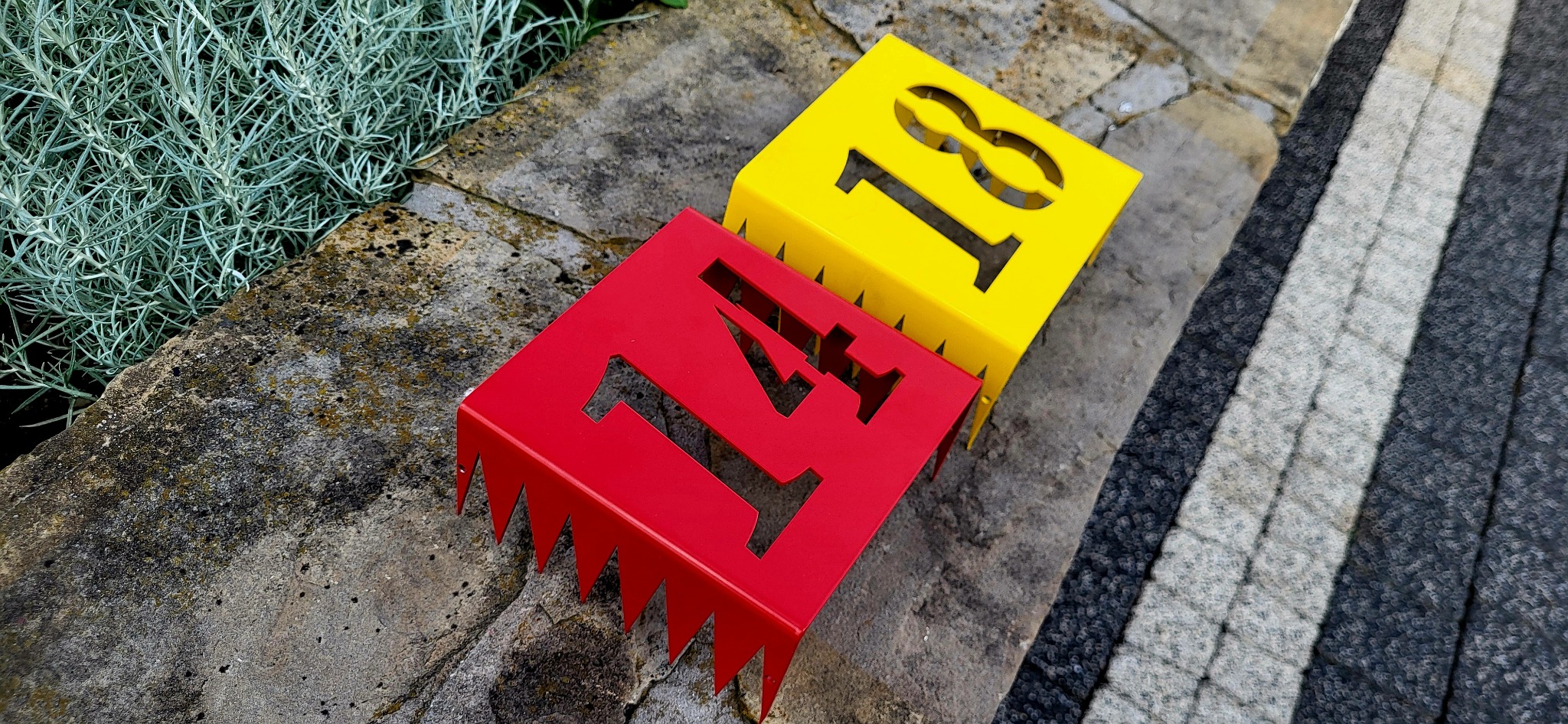 Czerwony i żółty, metalowy numer domu '17' i '18' z ząbkowanym dołem, leżą na kamiennej powierzchni obok roślinności. Wykonanie precyzyjne, nowoczesny design.