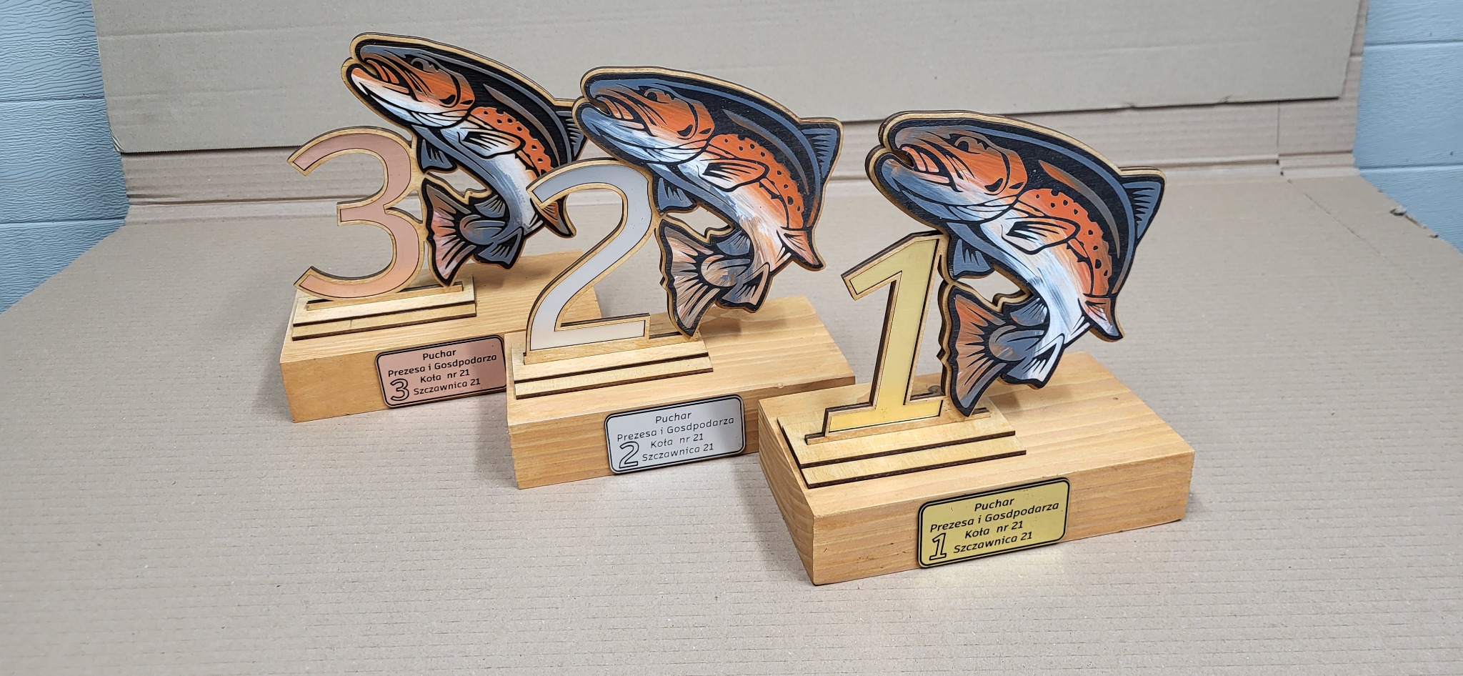 Ręcznie robione, drewniane trofea wędkarskie z numerami 1, 2 i 3, z motywem ryby, na jasnym tle. Unikalne nagrody dla zawodów wędkarskich.