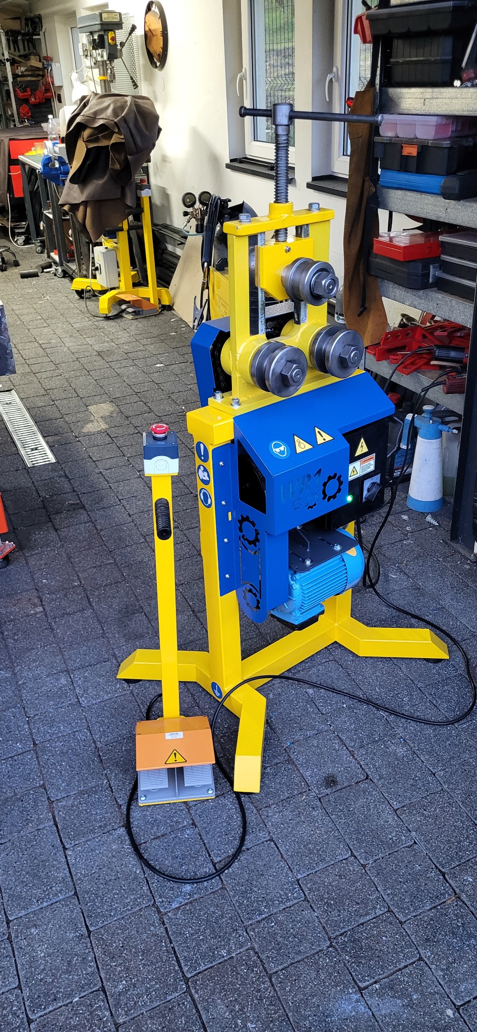 Giętarka trzypunktowa do rur i profili, pomalowana na żółto-niebiesko, z pedałem nożnym. Widoczne oznaczenia BHP i silnik elektryczny. Tło warsztatowe.