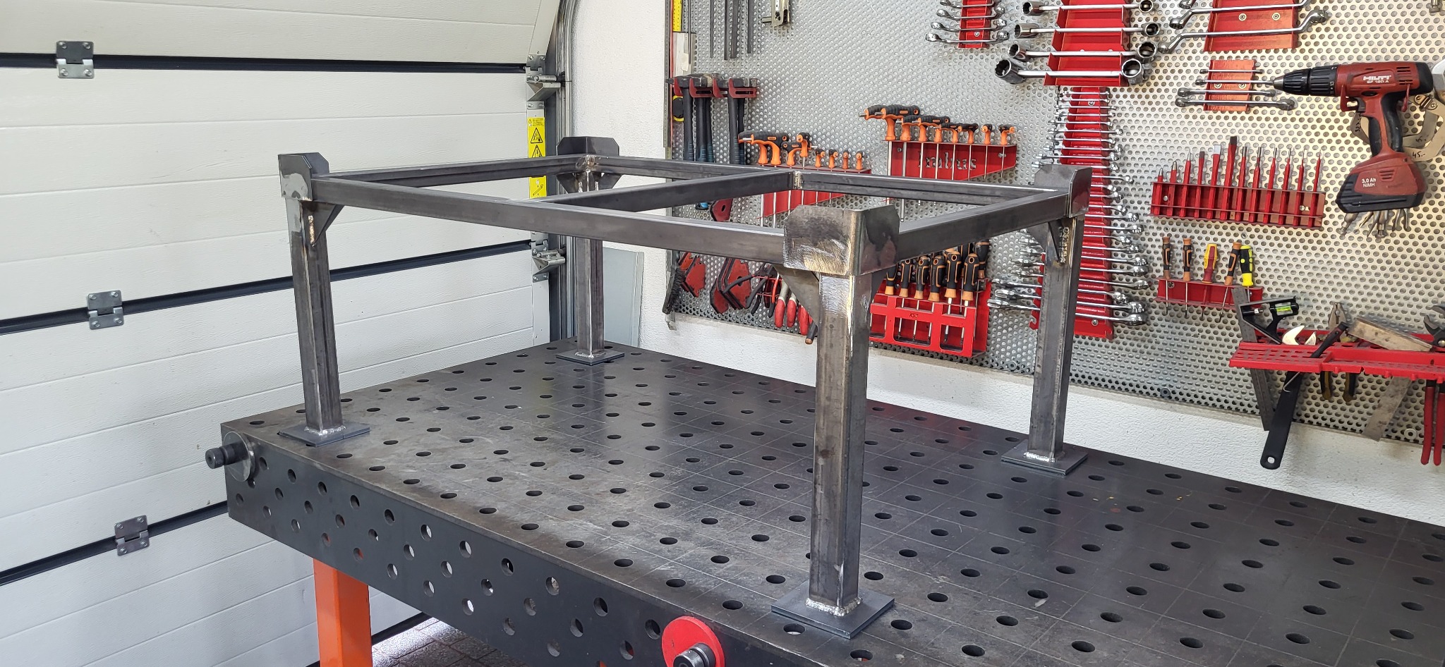 Metalowa konstrukcja stołu spawalniczego z profili kwadratowych, spawana, surowa, na tle ściany warsztatu z narzędziami. Widoczne spawy i perforowany blat stołu.