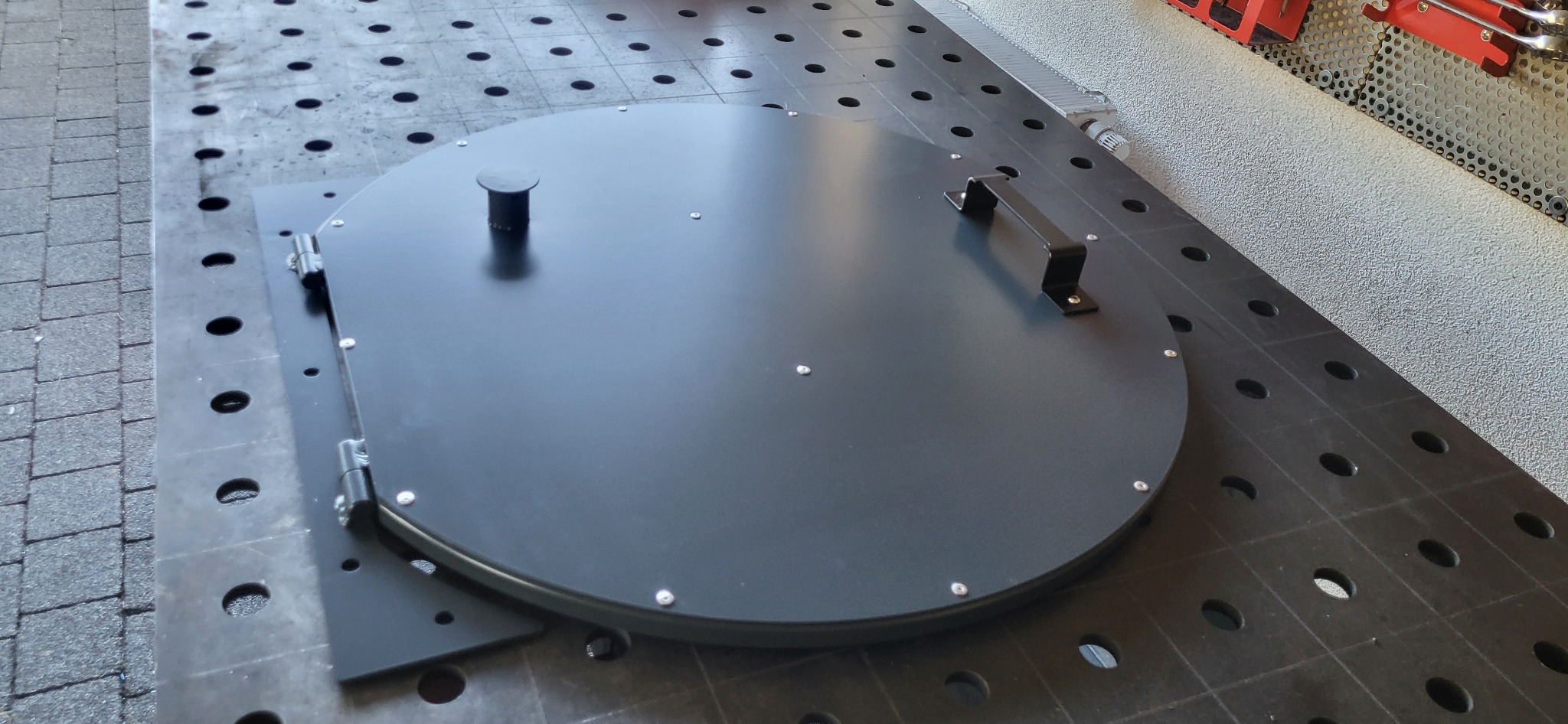 Czarna, okrągła pokrywa z uchwytem i zawiasami, przymocowana do perforowanego stołu warsztatowego. Widoczne nity i elementy mocujące. Detal wykonany z metalu.