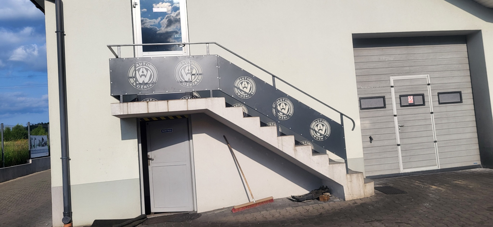 Schody zewnętrzne z metalową balustradą z wyciętym logo firmy Auto Woźniak, prowadzące do wejścia do budynku. Obok brama garażowa i drzwi do szatni.
