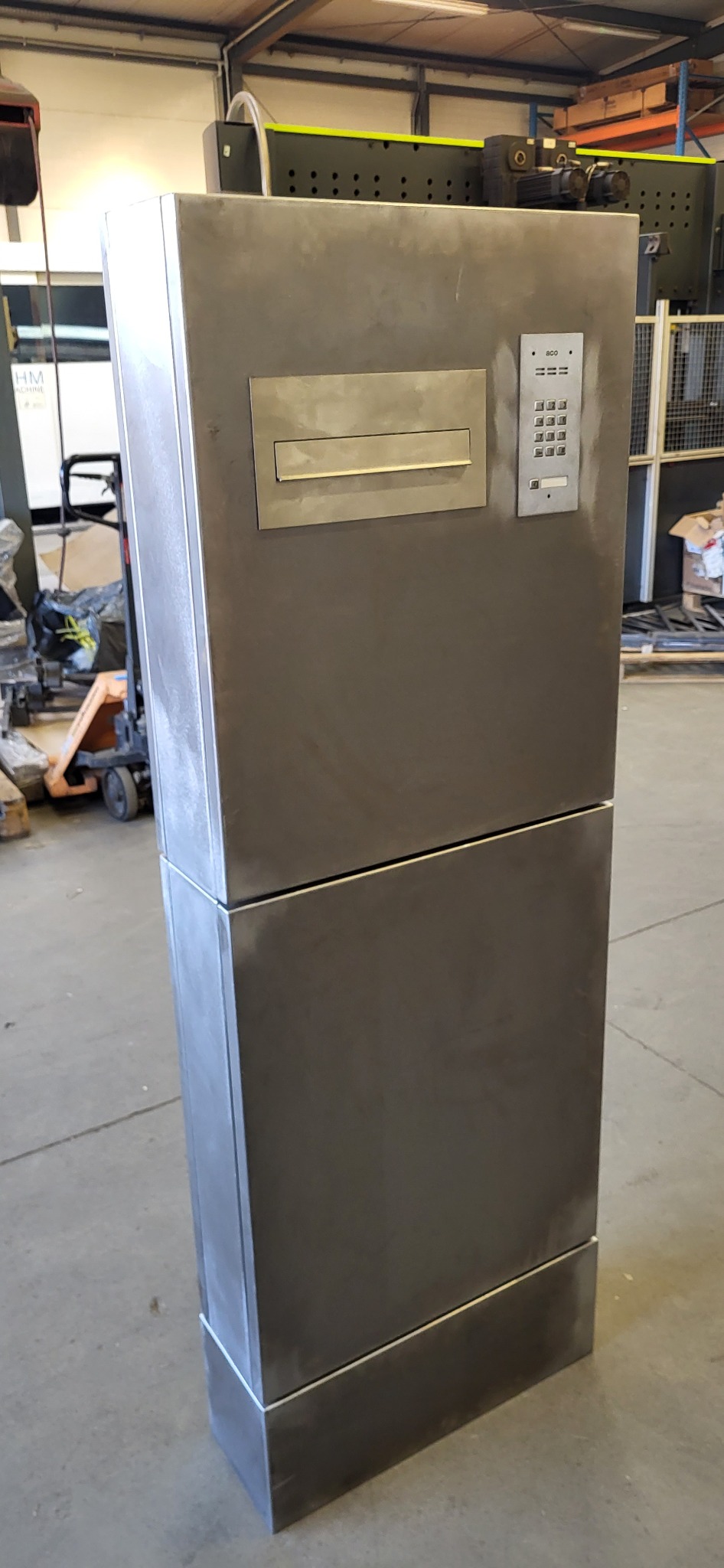 Metalowa skrzynka z panelem numerycznym i otworem na listy, wykonana z surowego metalu, stojąca w hali produkcyjnej. Widoczne ślady obróbki.