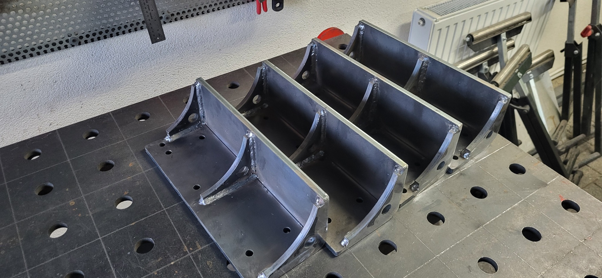 Cztery identyczne, spawane konstrukcje stalowe z otworami, ułożone rzędem na perforowanym stole warsztatowym. Widoczne spawy i surowy metal.