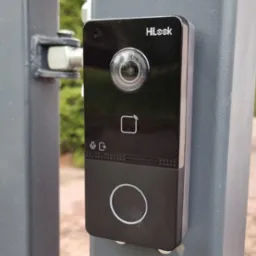 Czarny wideodomofon HiLook zamontowany na szarym słupku ogrodzenia, widok z bliska. Widoczna kamera, czytnik kart, mikrofon i przycisk dzwonka.