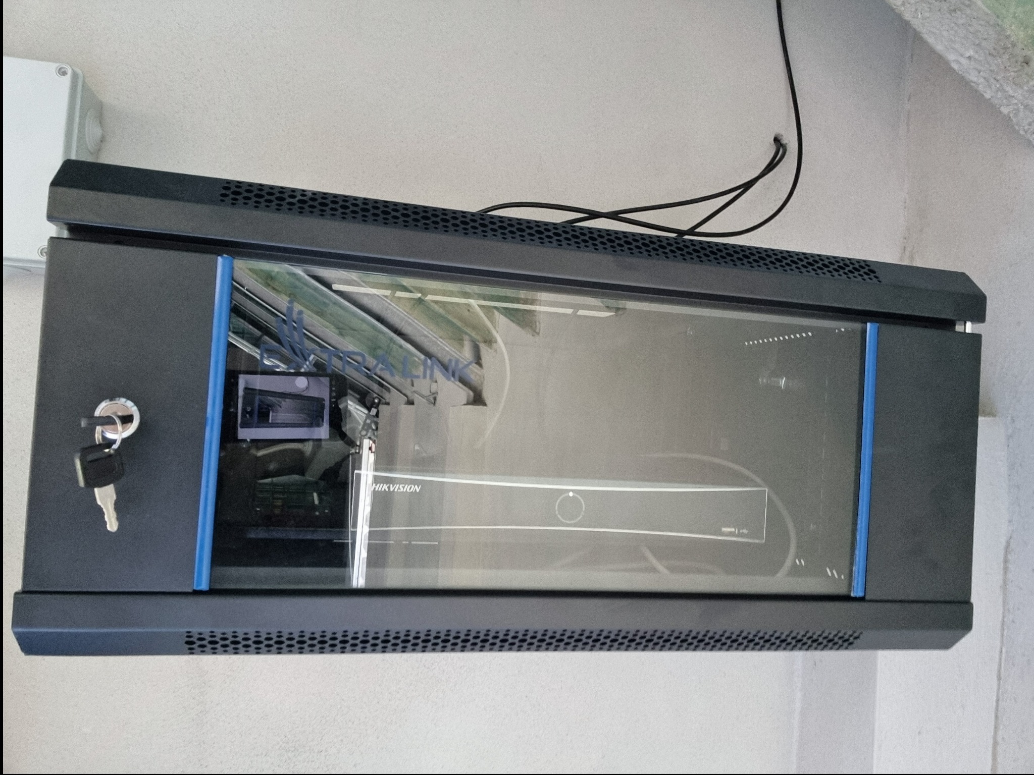 Szafa rack z zamkiem i kluczem, zawierająca rejestrator Hikvision, zamontowana na ścianie w Miechowie. Widoczne przewody i puszka instalacyjna.