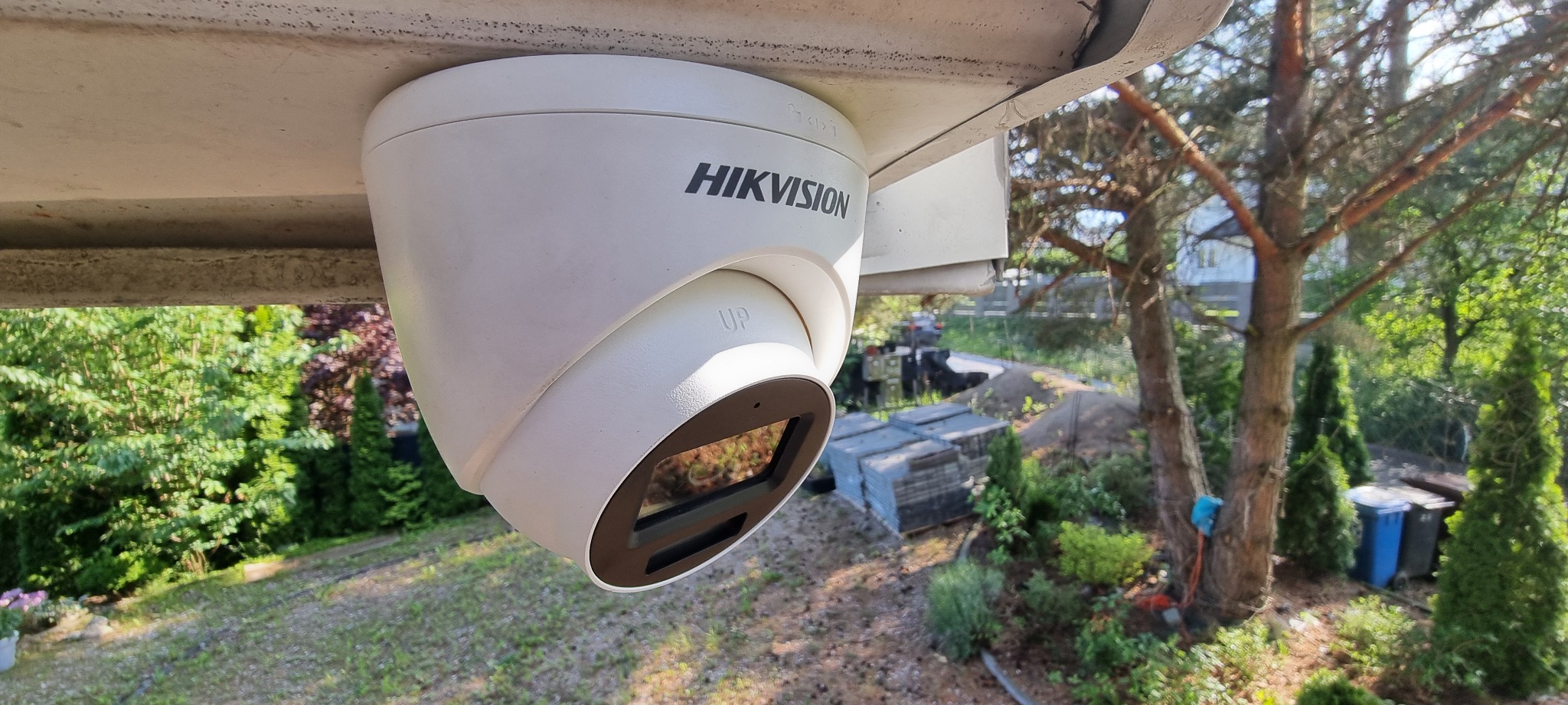 Kamera monitoringu Hikvision, zamontowana pod okapem, z widokiem na zieleń ogrodu. Obiektyw skierowany w dół, widoczne logo i napis 'UP'.
