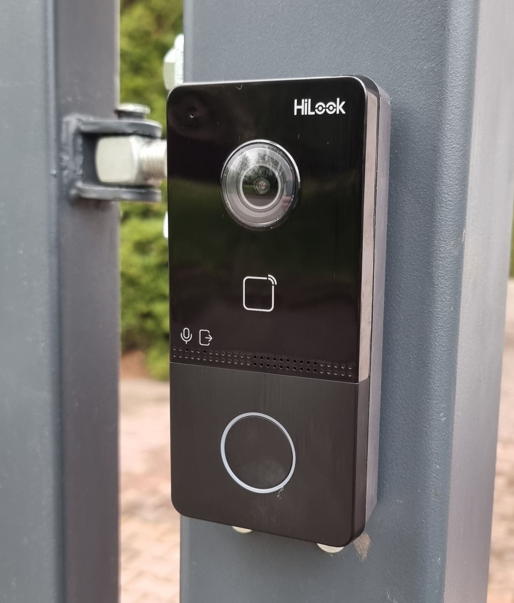 Czarny wideodomofon HiLook zamontowany na szarym słupku ogrodzenia, widok z bliska. Widoczna kamera, czytnik kart, mikrofon i przycisk dzwonka.