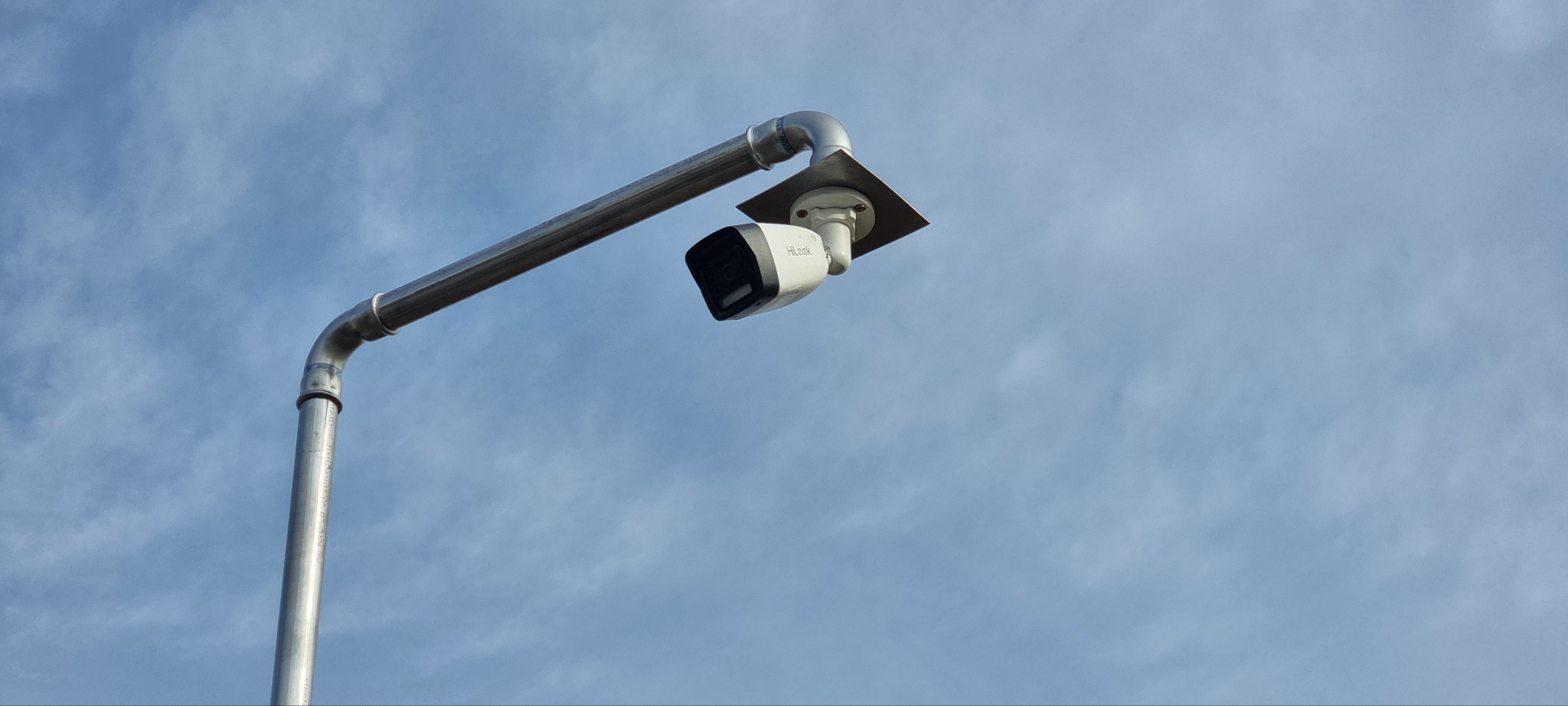 Kamera monitoringu HiLook na metalowym słupku, skierowana w dół, na tle błękitnego nieba. Widok z dołu, podkreślający solidność instalacji.