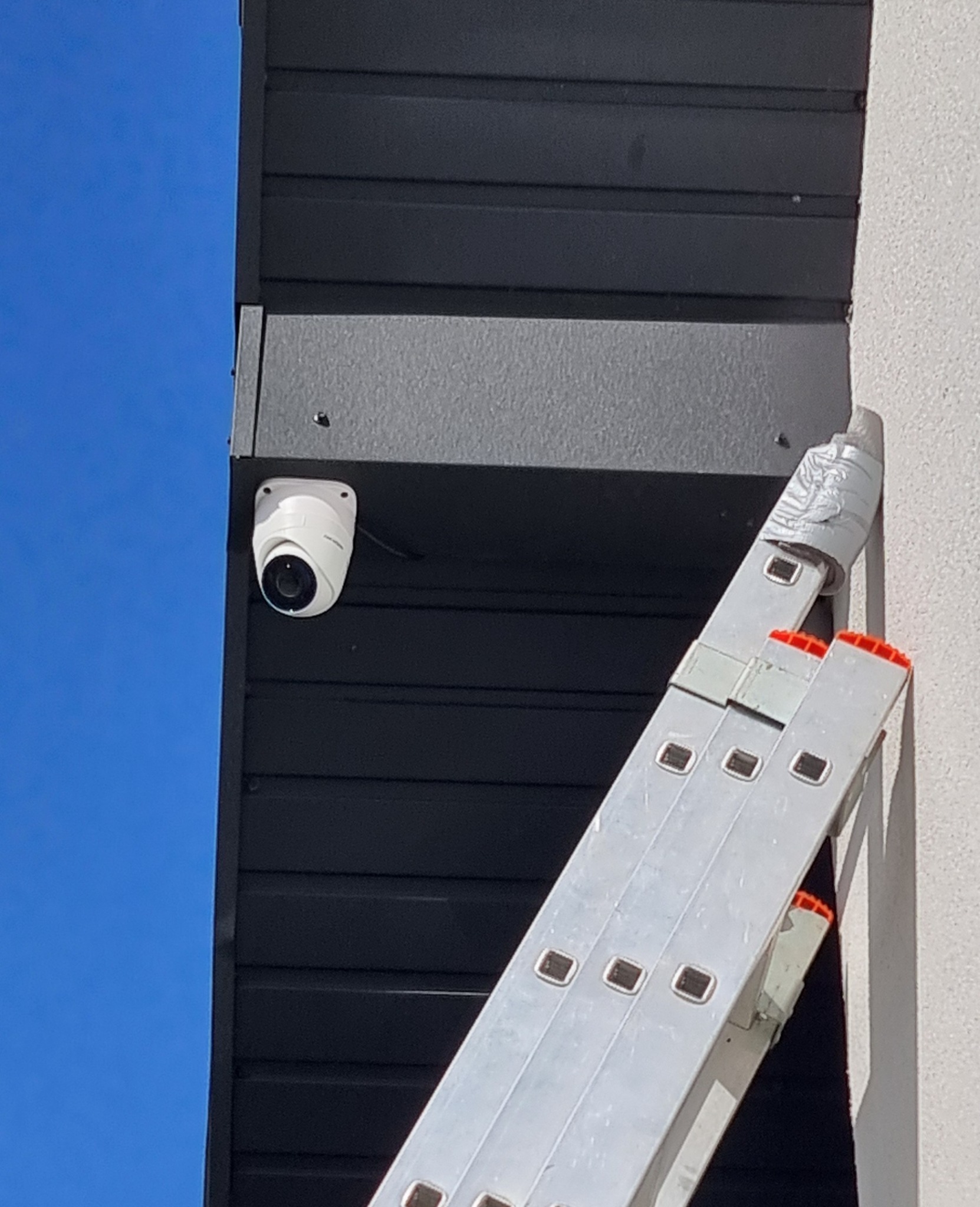 Kamera monitoringu zamontowana na elewacji budynku, widoczna drabina oparta o ścianę. Ujęcie z dołu, błękitne niebo w tle.