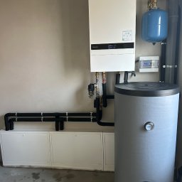 MG Instalacje Michał Górnowicz - Nowoczesny, biały piec gazowy Panasonic z szarym zasobnikiem wody i niebieskim naczyniem przeponowym, zamontowany na ścianie w pomieszczeniu technicznym. Widoczne rury i instalacja.