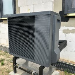 MG Instalacje Michał Górnowicz - Szara pompa ciepła Panasonic Aquarea na zewnątrz budynku z białego bloczka, zamontowana na metalowej konstrukcji. Widoczne okno i fragment ściany.