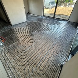 MG Instalacje Michał Górnowicz - Ułożone rury ogrzewania podłogowego na folii izolacyjnej w jasnym pomieszczeniu z dużym oknem, przez które widać zaparkowany samochód dostawczy.