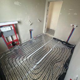 MG Instalacje Michał Górnowicz - Ułożone rury ogrzewania podłogowego na folii aluminiowej, widoczne przyłącza do ściany i stelaż podtynkowy WC TECE. Surowe ściany w trakcie prac wykończeniowych.
