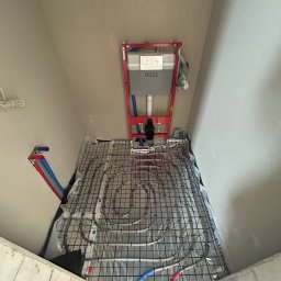 MG Instalacje Michał Górnowicz - Montaż ogrzewania podłogowego w łazience z widocznym stelażem WC TECE. Rury grzewcze ułożone na folii izolacyjnej, gotowe do zalania wylewką. Ujęcie z góry.