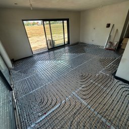 MG Instalacje Michał Górnowicz - Ułożone rury ogrzewania podłogowego na folii izolacyjnej w jasnym pomieszczeniu z dużym oknem tarasowym, widok z góry, etap instalacji przed wylewką.