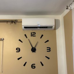 MG Instalacje Michał Górnowicz - Biały klimatyzator Kaisai zamontowany na beżowej ścianie nad dużym, czarnym zegarem ściennym. Widoczny fragment okna z zasłoną.