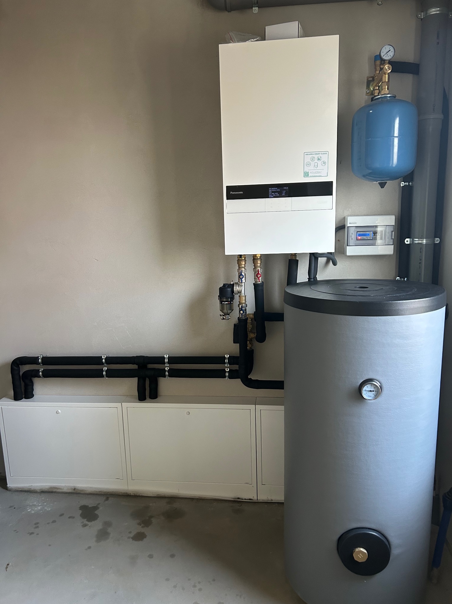 Nowoczesny, biały piec gazowy Panasonic z szarym zasobnikiem wody i niebieskim naczyniem przeponowym, zamontowany na ścianie w pomieszczeniu technicznym. Widoczne rury i instalacja.