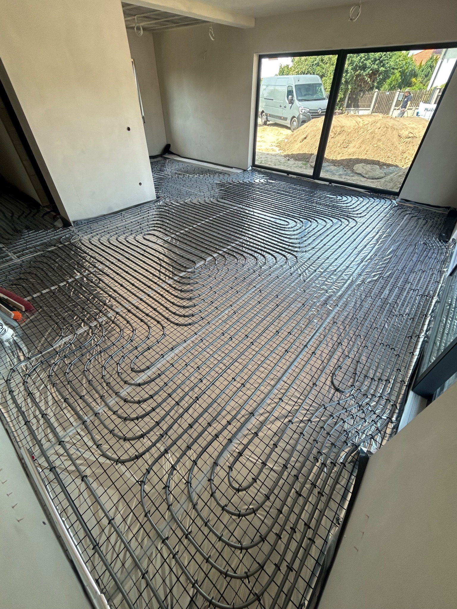 Ułożone rury ogrzewania podłogowego na folii izolacyjnej w jasnym pomieszczeniu z dużym oknem, przez które widać zaparkowany samochód dostawczy.