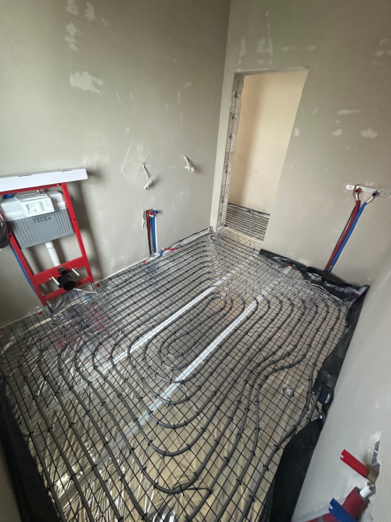 Ułożone rury ogrzewania podłogowego na folii aluminiowej, widoczne przyłącza do ściany i stelaż podtynkowy WC TECE. Surowe ściany w trakcie prac wykończeniowych.