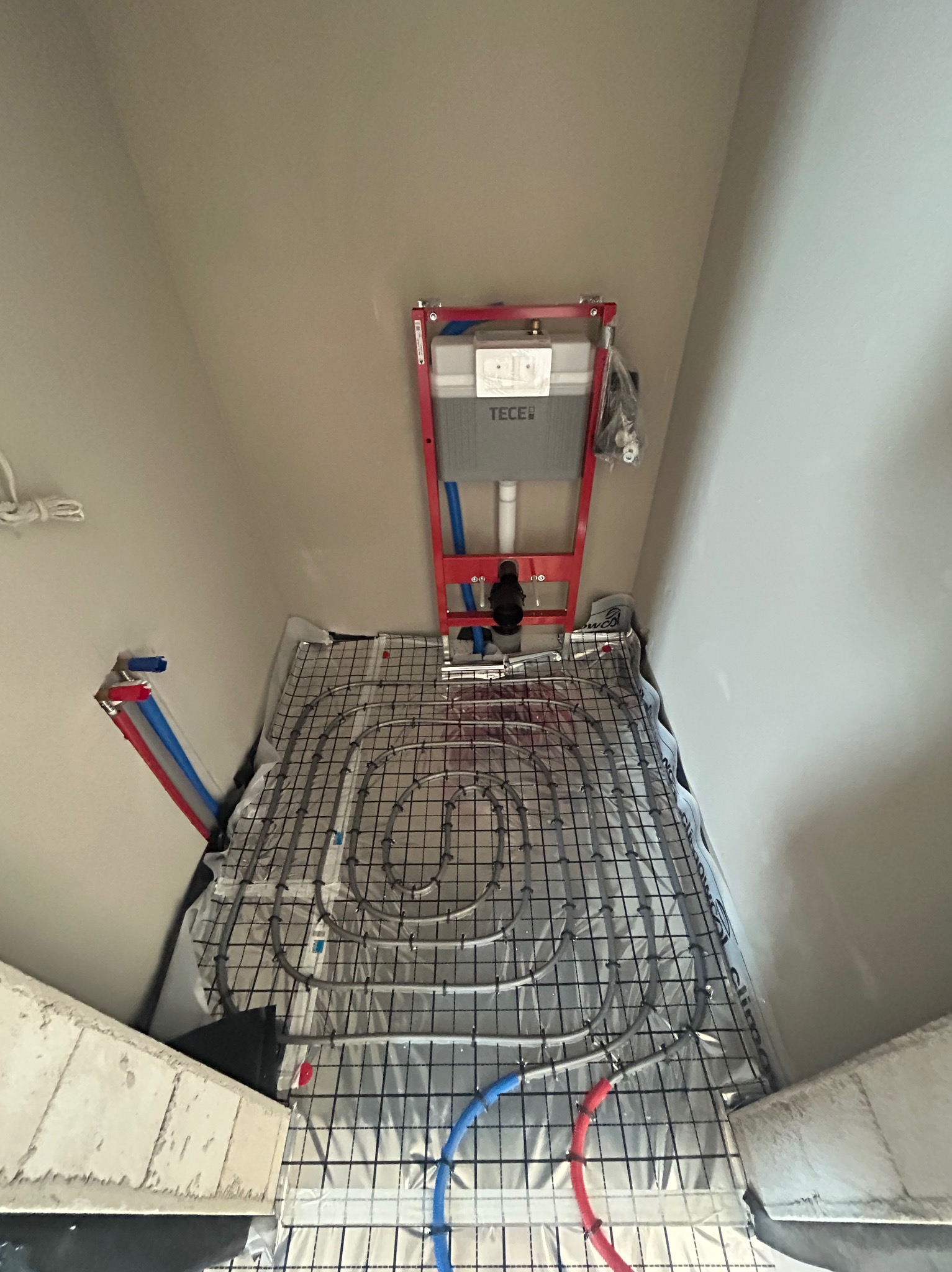 Montaż ogrzewania podłogowego w łazience z widocznym stelażem WC TECE. Rury grzewcze ułożone na folii izolacyjnej, gotowe do zalania wylewką. Ujęcie z góry.