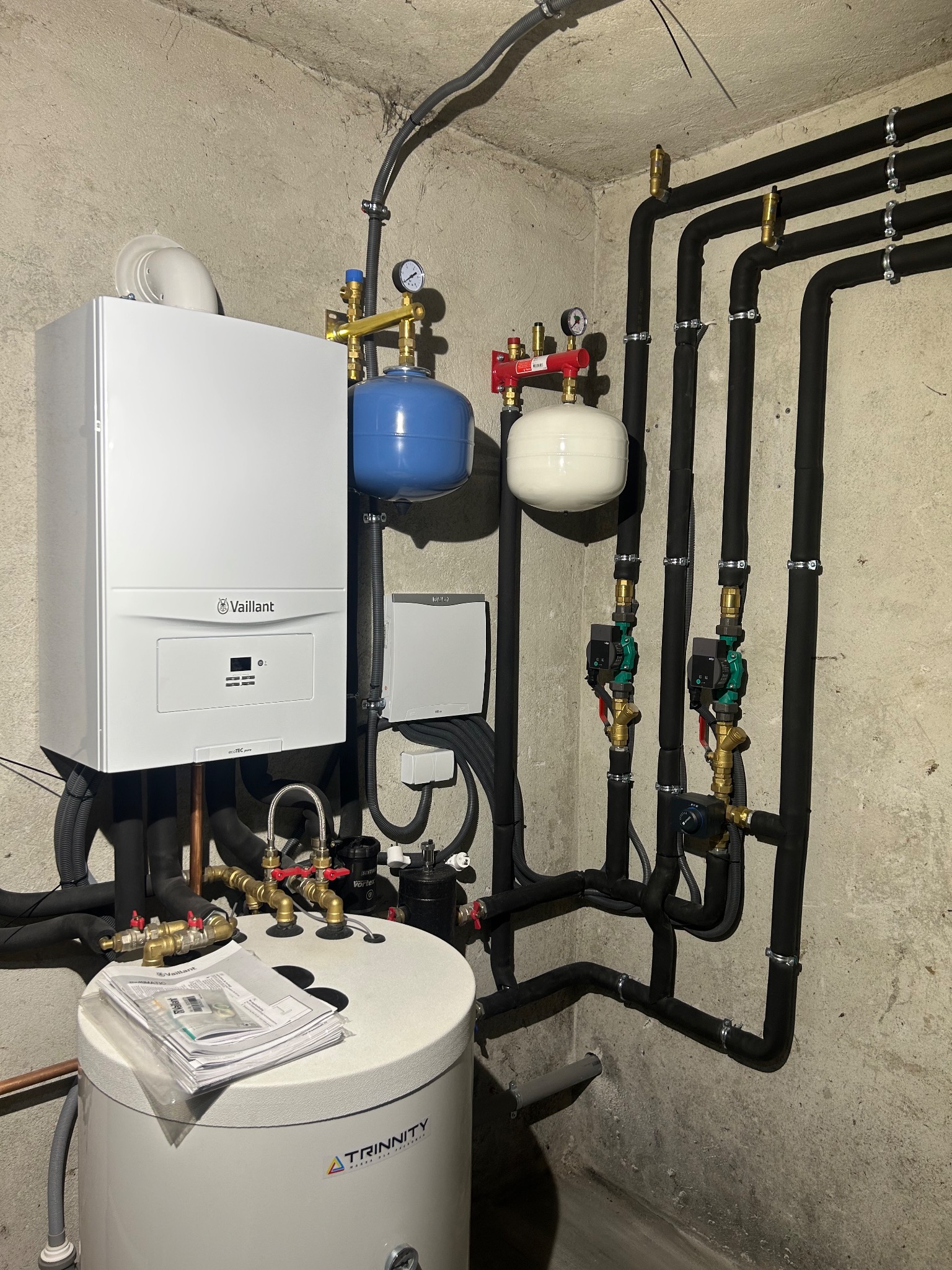 Nowoczesna kotłownia z piecem Vaillant, zbiornikiem Trinity i czarnymi rurami w izolacji. Widoczne manometry i zawory. Instalacja w surowym pomieszczeniu.