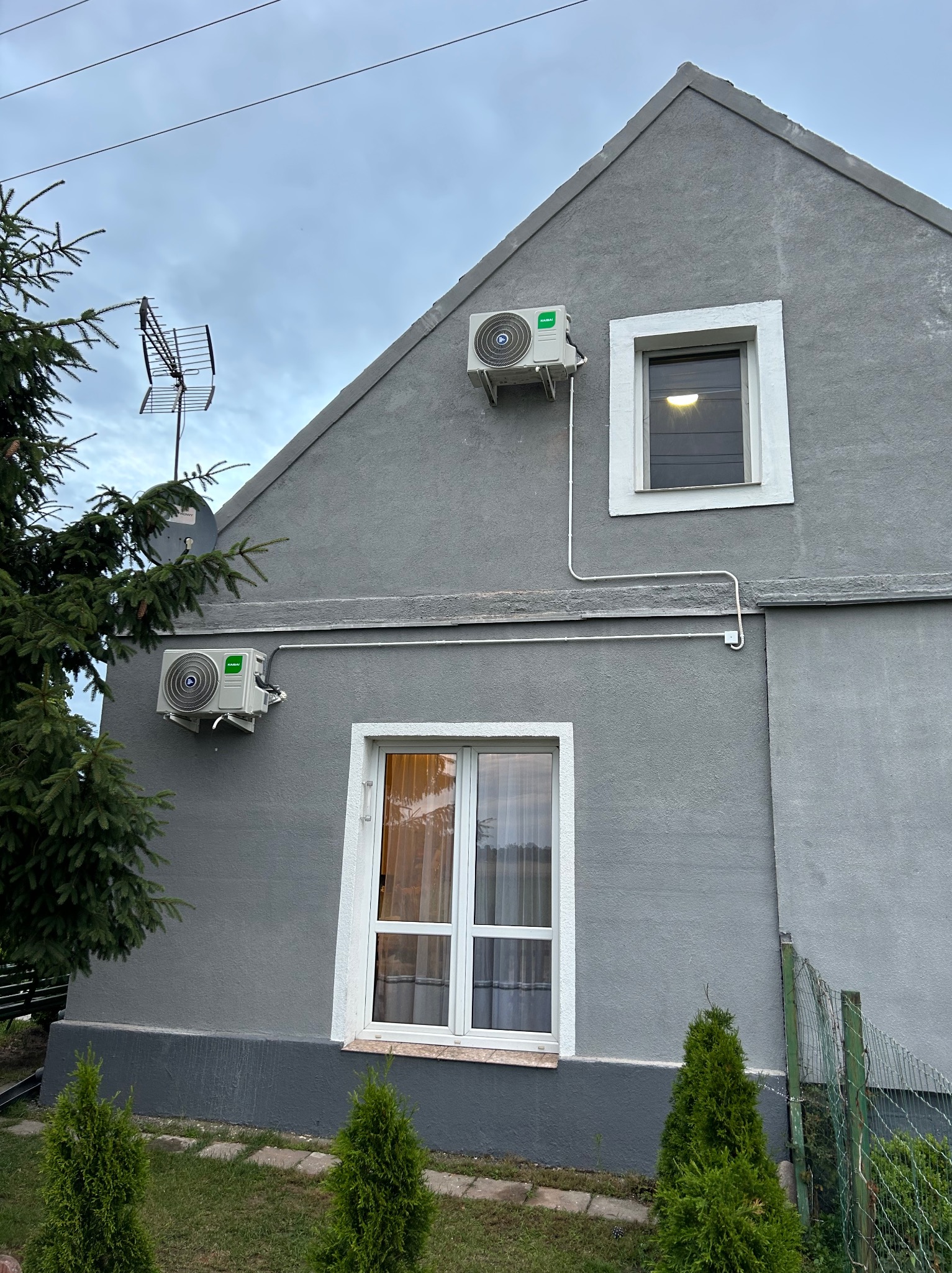 Szary dom z dwoma klimatyzatorami na elewacji, widoczne białe rury instalacyjne, okna z białymi ramami i fragment zieleni. Ujęcie z dołu, pochmurne niebo.