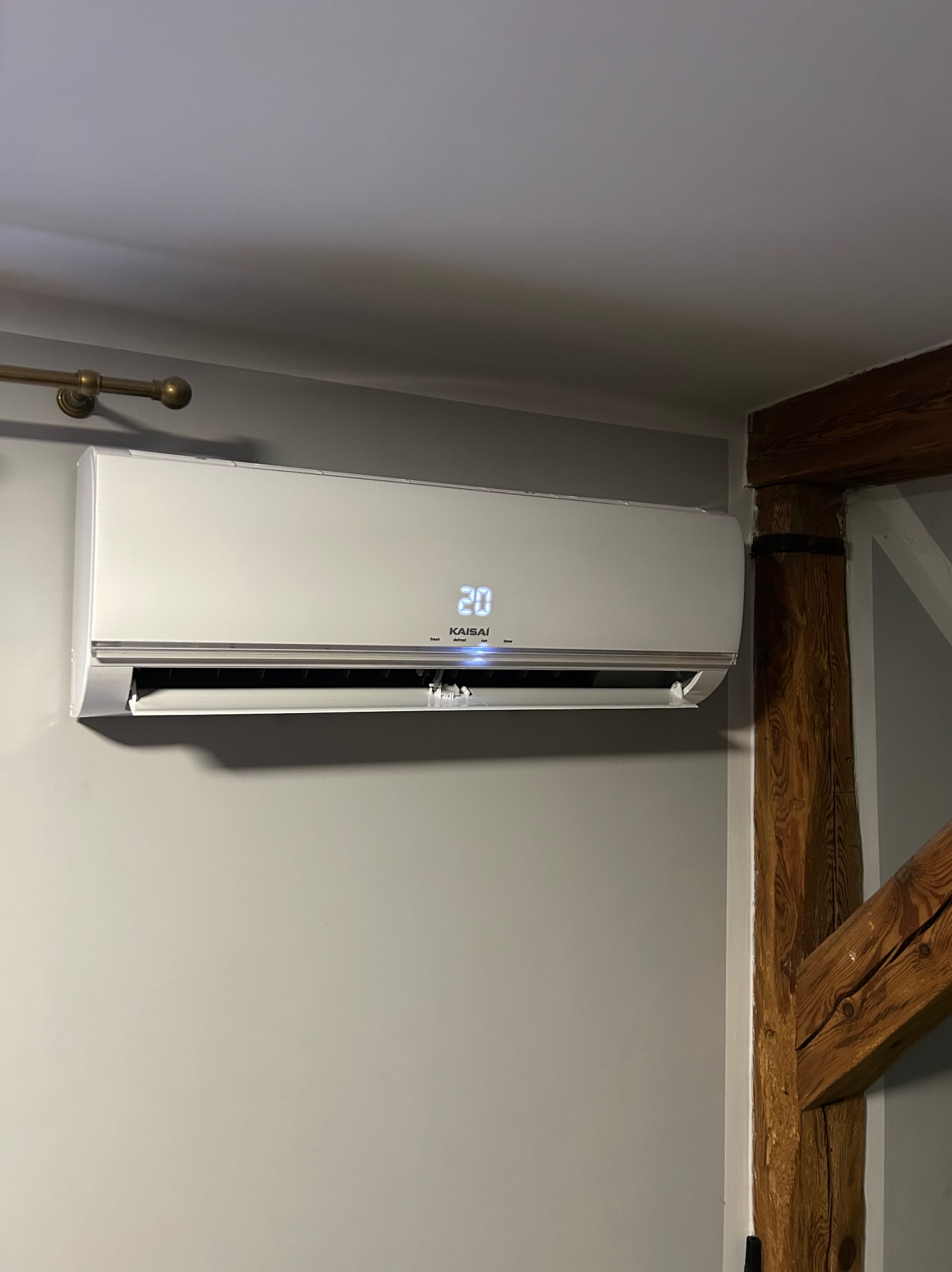 Biały klimatyzator Kaisai na szarej ścianie, obok drewnianej belki stropowej. Wyświetlacz pokazuje 20 stopni. Widoczne elementy instalacji.