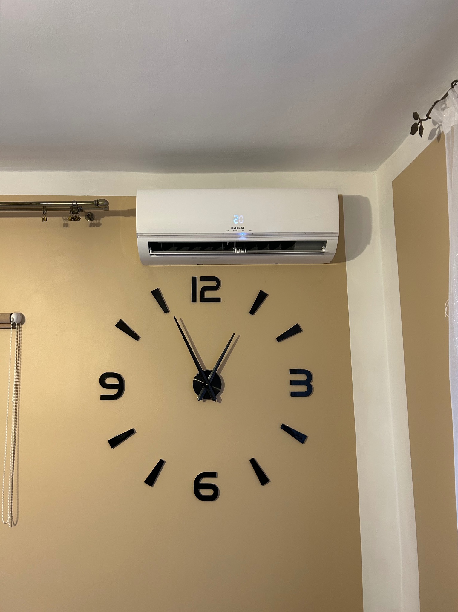 Biały klimatyzator Kaisai zamontowany na beżowej ścianie nad dużym, czarnym zegarem ściennym. Widoczny fragment okna z zasłoną.
