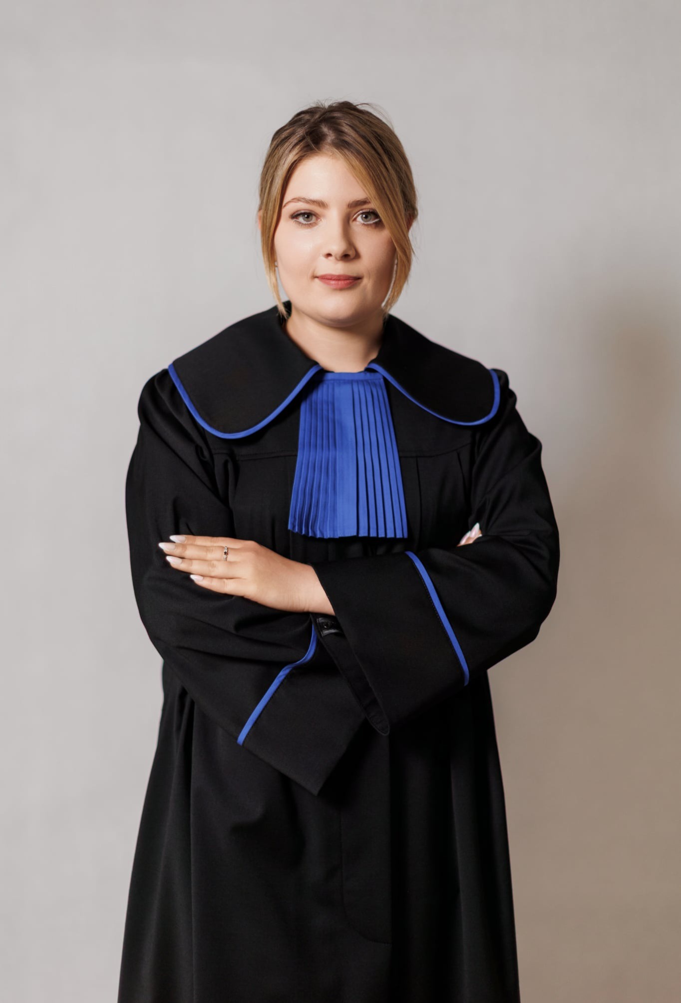 Portret kobiety-adwokata w todze z niebieskimi akcentami, stojącej na neutralnym tle z założonymi rękami, prezentującej profesjonalizm i pewność siebie.