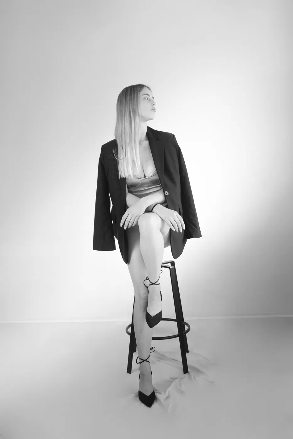 Czarno-biała sesja kobieca: blondynka w marynarce i szpilkach siedzi na stołku, spoglądając w bok. Minimalistyczne tło, elegancka poza, podkreślona sylwetka.