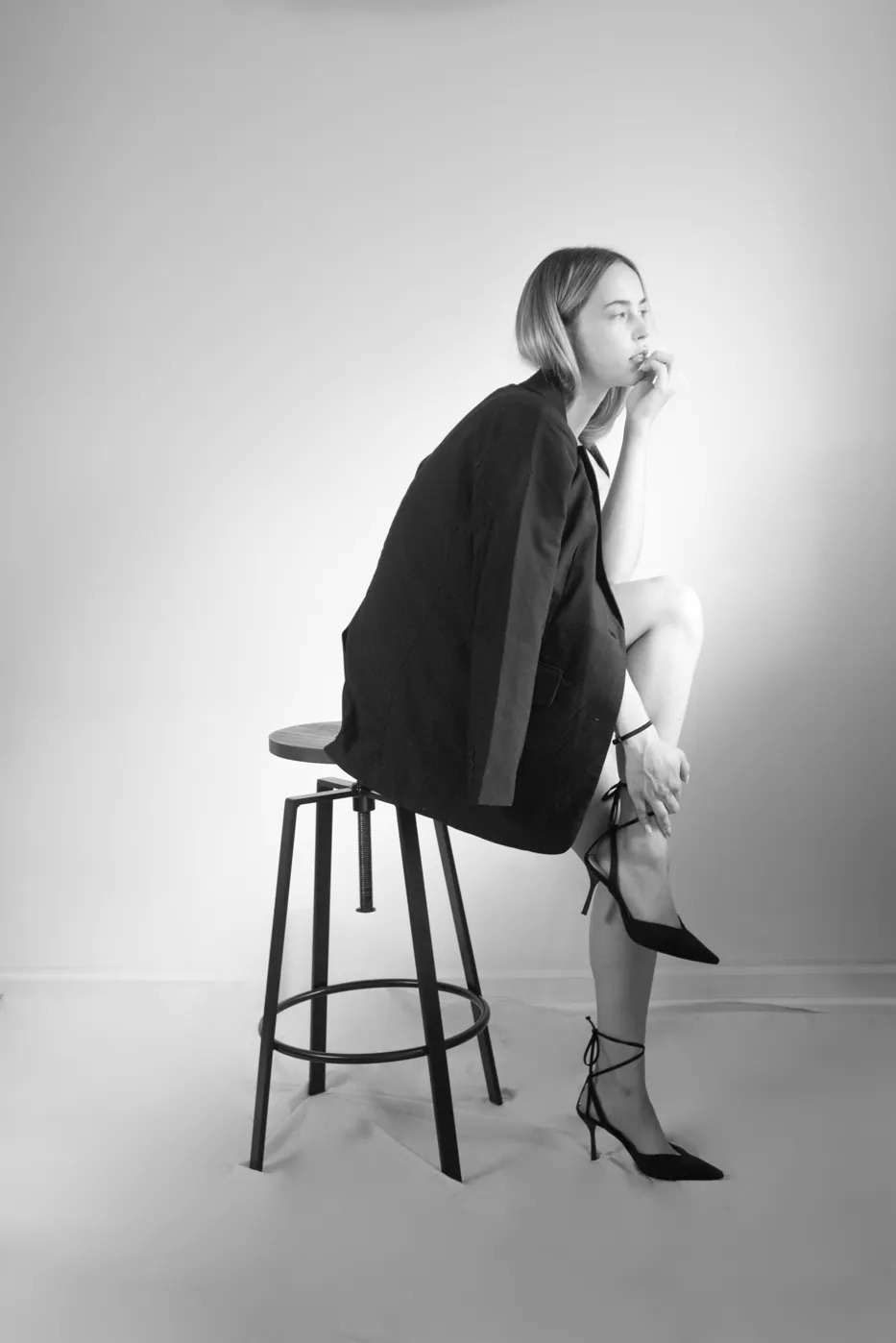 Czarno-białe zdjęcie kobiety siedzącej na stołku w marynarce i szpilkach, zamyślona poza, minimalistyczne tło, oświetlenie podkreśla kontury sylwetki.