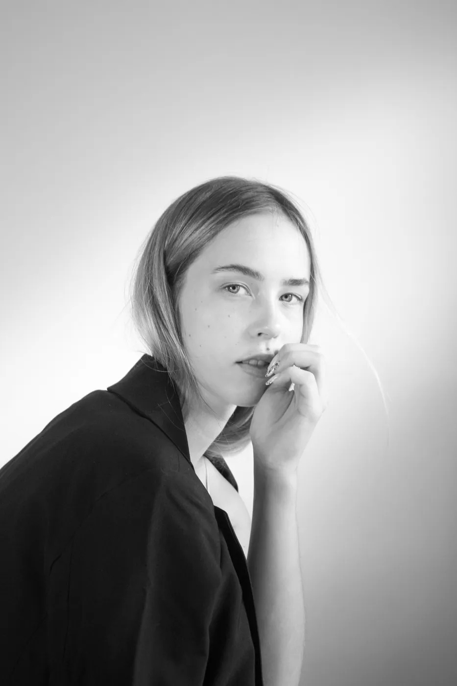 Portret młodej kobiety w czarnej marynarce, z dłonią przy ustach, w czarno-białej tonacji. Delikatne światło podkreśla rysy twarzy i subtelny makijaż.