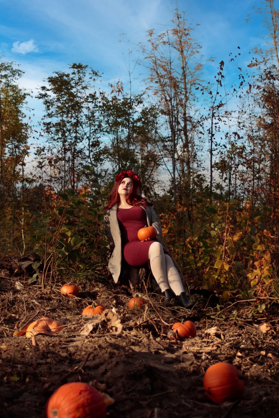 Jesienna sesja w plenerze: Kobieta w bordowej sukience i wianku z kwiatów siedzi na pniu drzewa, otoczona dyniami na tle lasu z błękitnym niebem w tle.