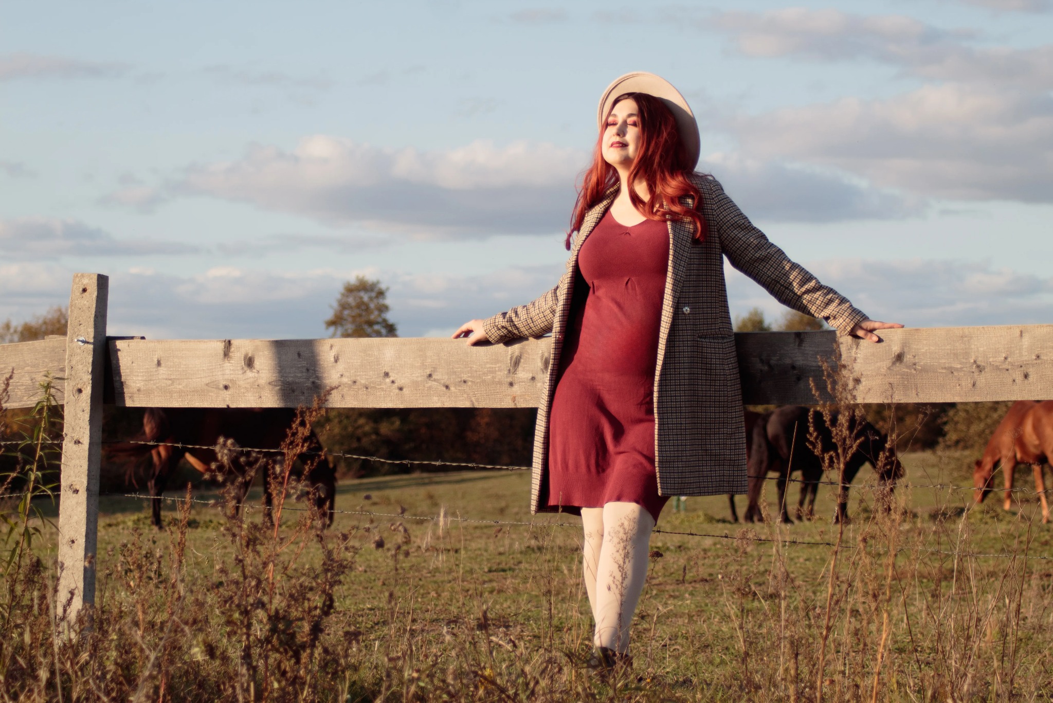 Kobieta w kapeluszu i płaszczu opiera się o drewniany płot na tle pastwiska z końmi. Ciepłe światło zachodzącego słońca. Ujęcie w plenerze, stylizowane.