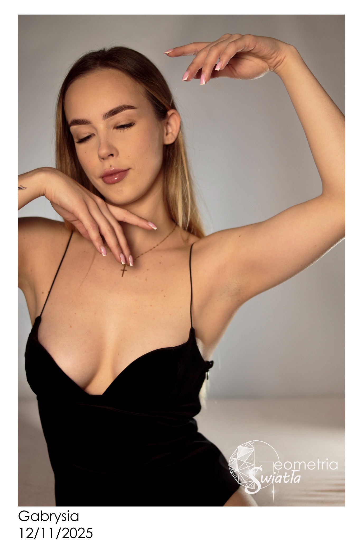 Portret kobiety w czarnej sukience na ramiączkach, z delikatnym makijażem i złotym naszyjnikiem z krzyżykiem, pozującej na jasnym tle. Widoczne logo fotografa.