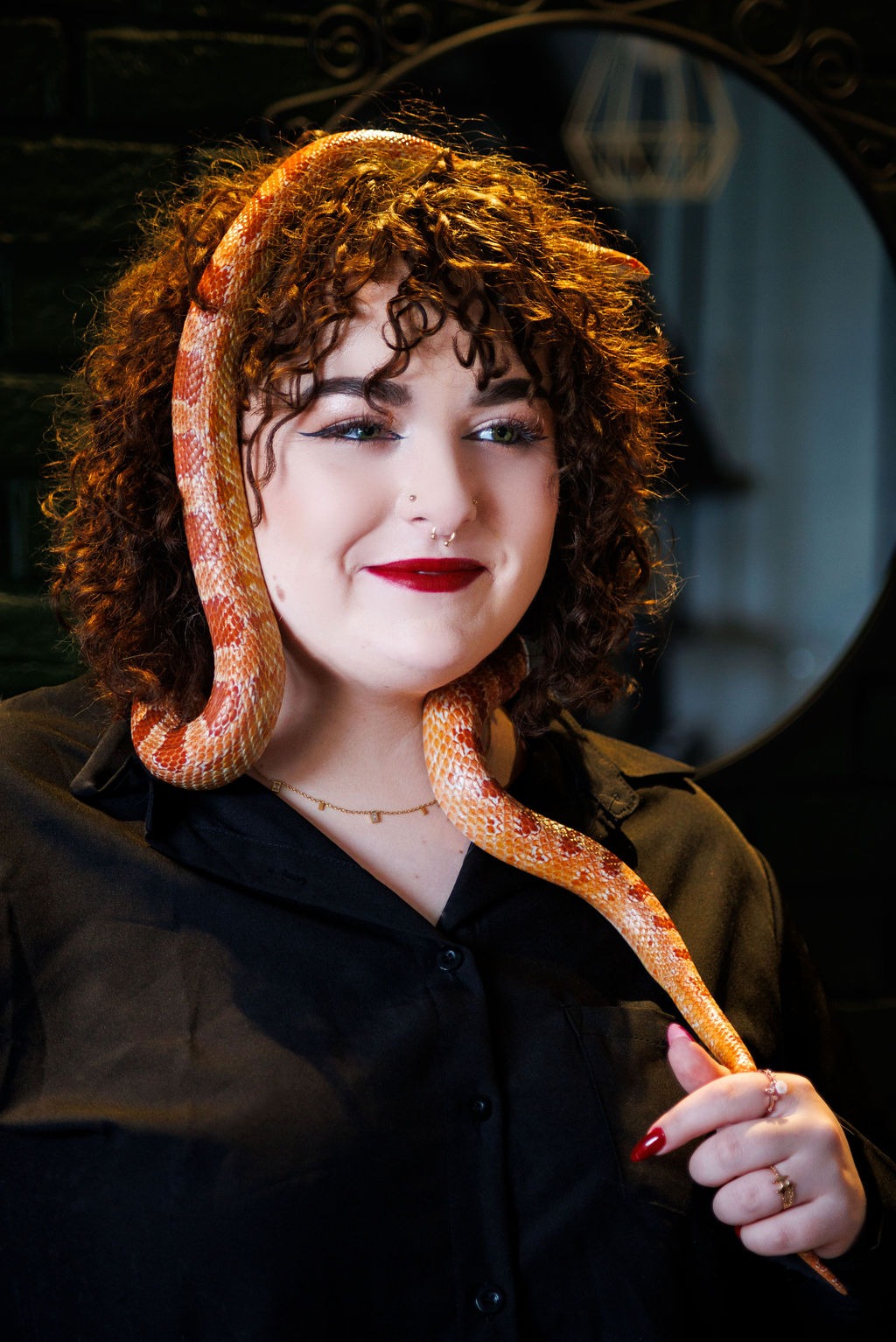 Portret kobiety z kręconymi włosami i w czarnej koszuli, na której szyi i głowie owinięty jest pomarańczowy wąż. Kobieta ma czerwone usta i piercing w nosie.