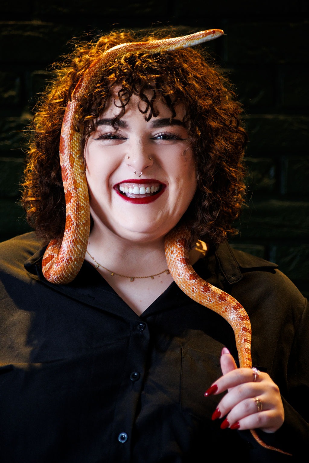 Portret uśmiechniętej kobiety z rudymi kręconymi włosami i wężem zbożowym owiniętym wokół szyi i głowy, na ciemnym tle. Wyraziste czerwone usta i ciemna koszula.