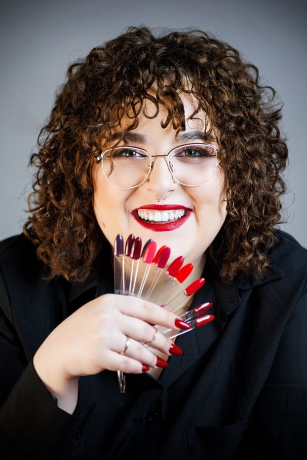 Uśmiechnięta brunetka w okularach prezentuje wachlarz kolorów lakierów do paznokci, z dopasowanym czerwonym manicure i makijażem ust. Szare tło.