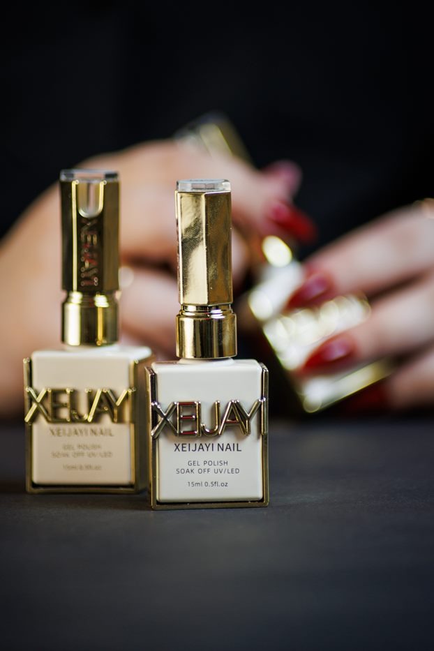 Dwa flakony lakieru hybrydowego Xeijayi Nail z złotymi elementami na ciemnym tle, w tle dłonie z czerwonym manicure. Produkt do stylizacji paznokci.