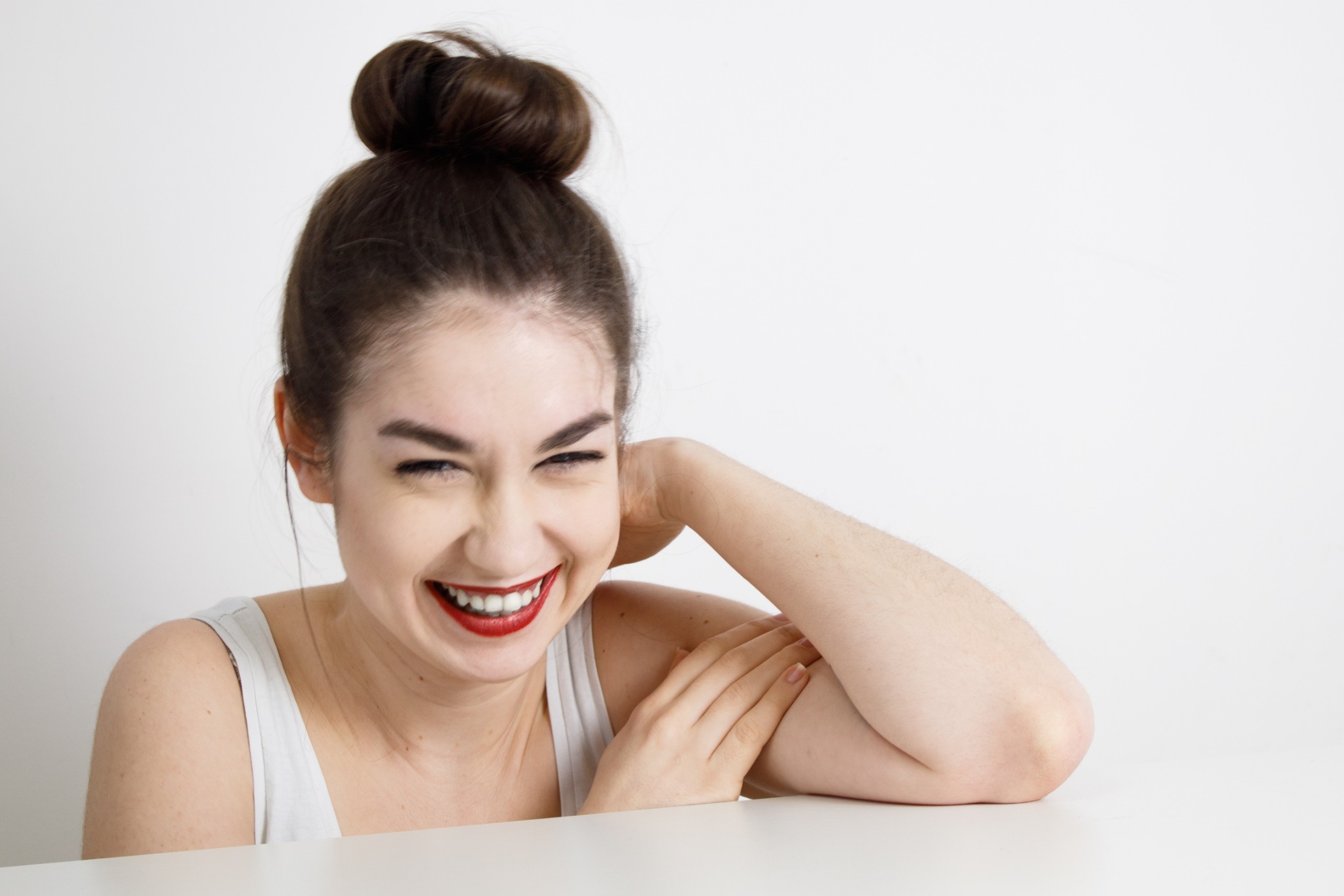 Portret uśmiechniętej brunetki z włosami upiętymi w kok, opierającej się o biały stół. Intensywna czerwień ust i radosny wyraz twarzy na jasnym tle.