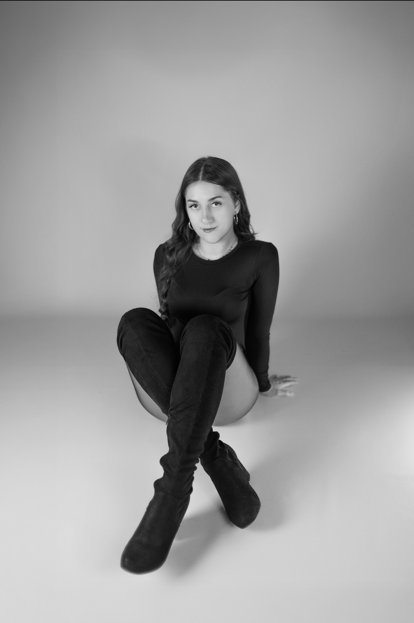 Monochromatyczne, pełne ujęcie kobiety w body i kozakach za kolano, siedzącej na jasnym tle. Delikatne światło podkreśla jej rysy twarzy i długie włosy.