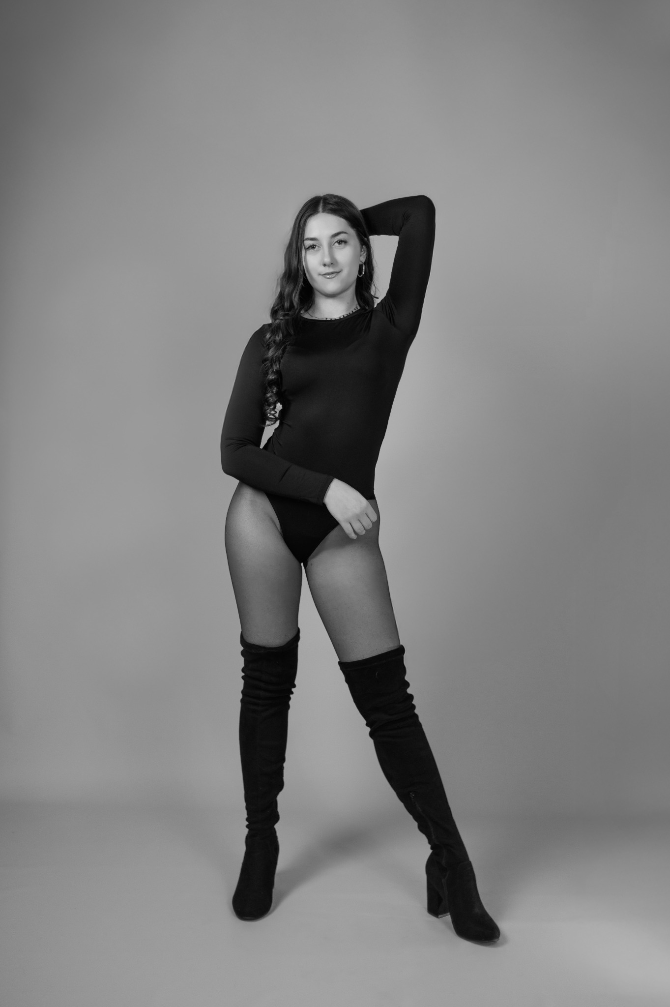 Czarno-białe, pełne ujęcie kobiety w body i kozakach za kolano, pozującej na szarym tle. Ręka uniesiona nad głową, włosy splecione w warkocz. Minimalistyczny styl.