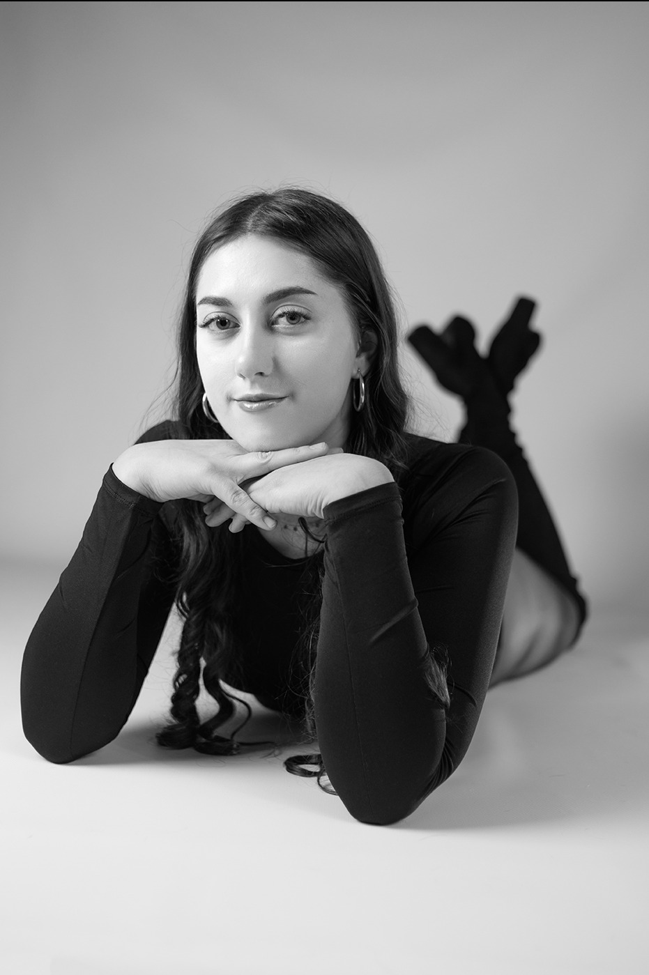 Czarno-białe zdjęcie kobiety leżącej na brzuchu, opierającej brodę na dłoniach, z nogami uniesionymi do góry. Ubrana w ciemny strój, z kolczykami kołami. Gładkie tło.