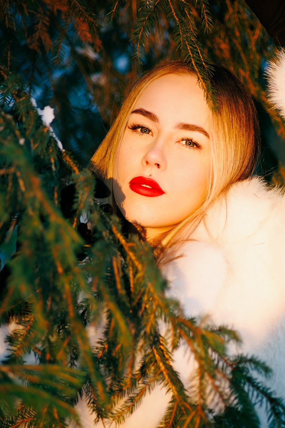 Portret kobiety z blond włosami i czerwonymi ustami, otoczonej gałązkami choinki w zimowej scenerii, oświetlonej ciepłym światłem.