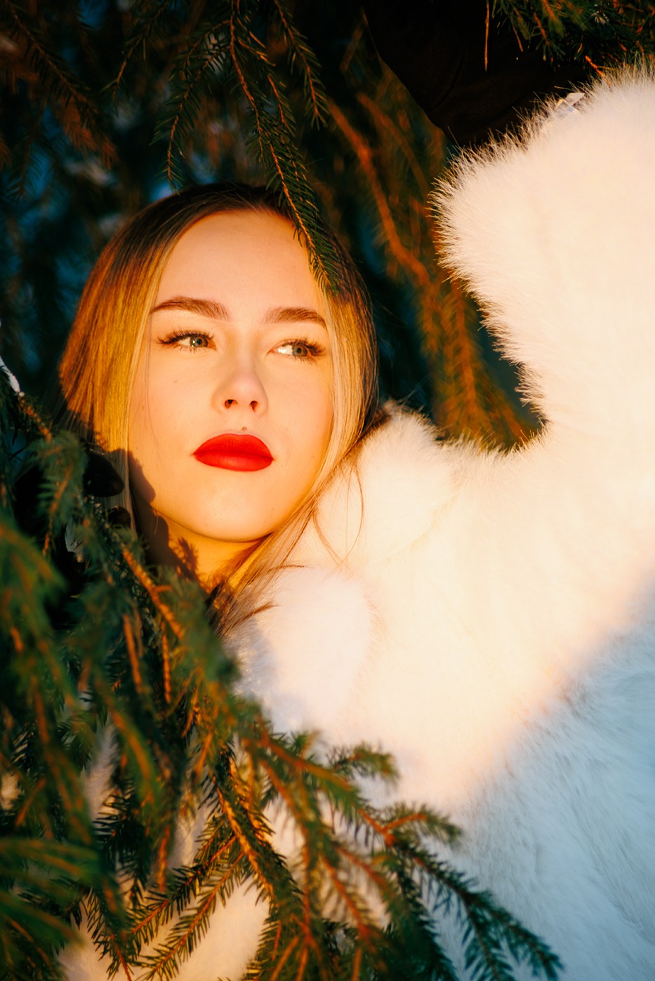 Portret kobiety w futrze, otoczonej gałązkami świerku, z mocnym makijażem ust w odcieniu czerwieni, oświetlonej ciepłym, naturalnym światłem.