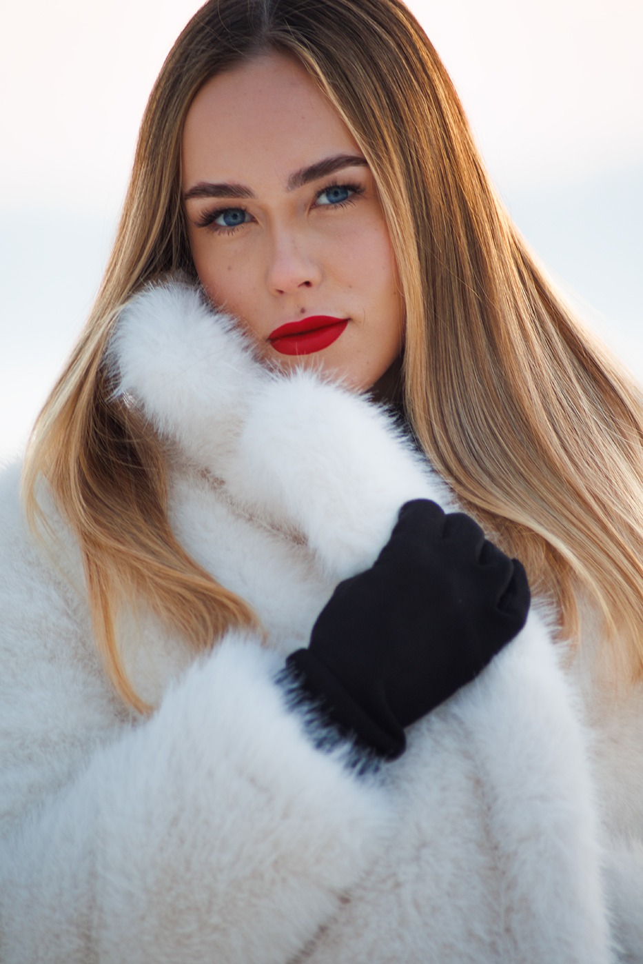 Portret kobiety o blond włosach i niebieskich oczach, z czerwoną szminką, w białym futrze i czarnych rękawiczkach, spoglądającej w obiektyw.